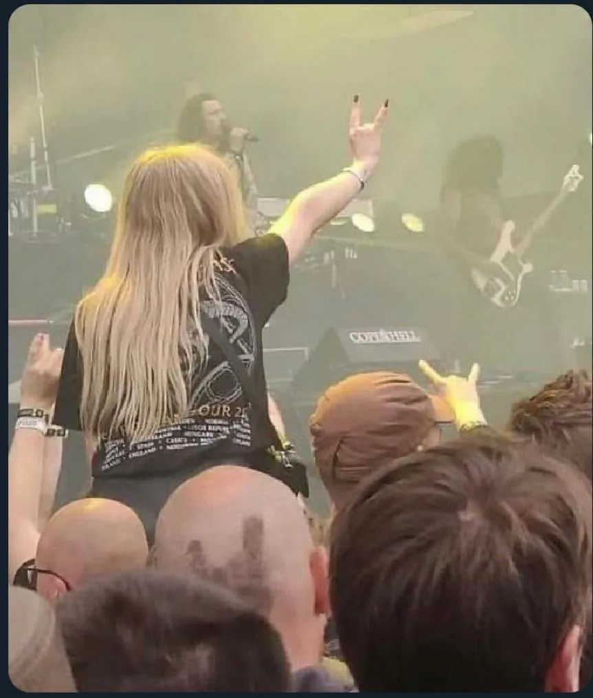 A person with long blonde hair, seen from behind, is at a concert wearing a black t-shirt with 'Metallica Tour 2024' printed…