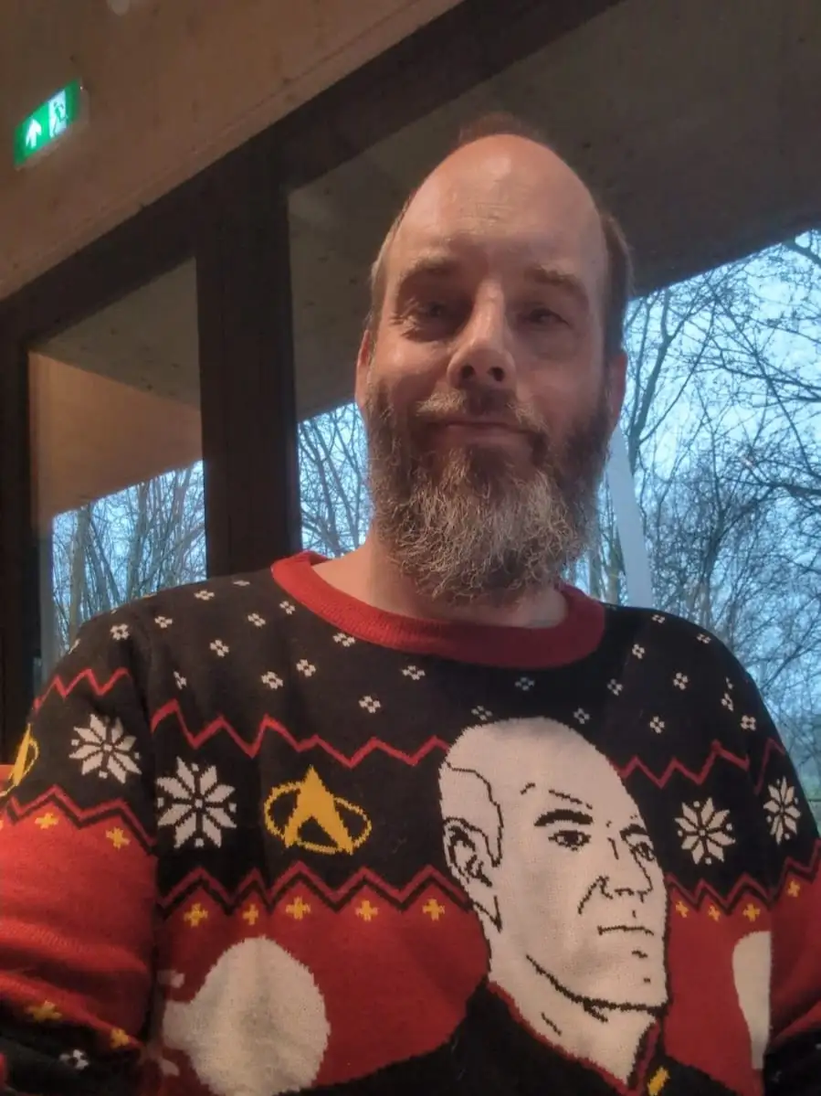 a man with a beard and receding hairline wears a festive black, red, and white Christmas sweater featuring a large portrait…