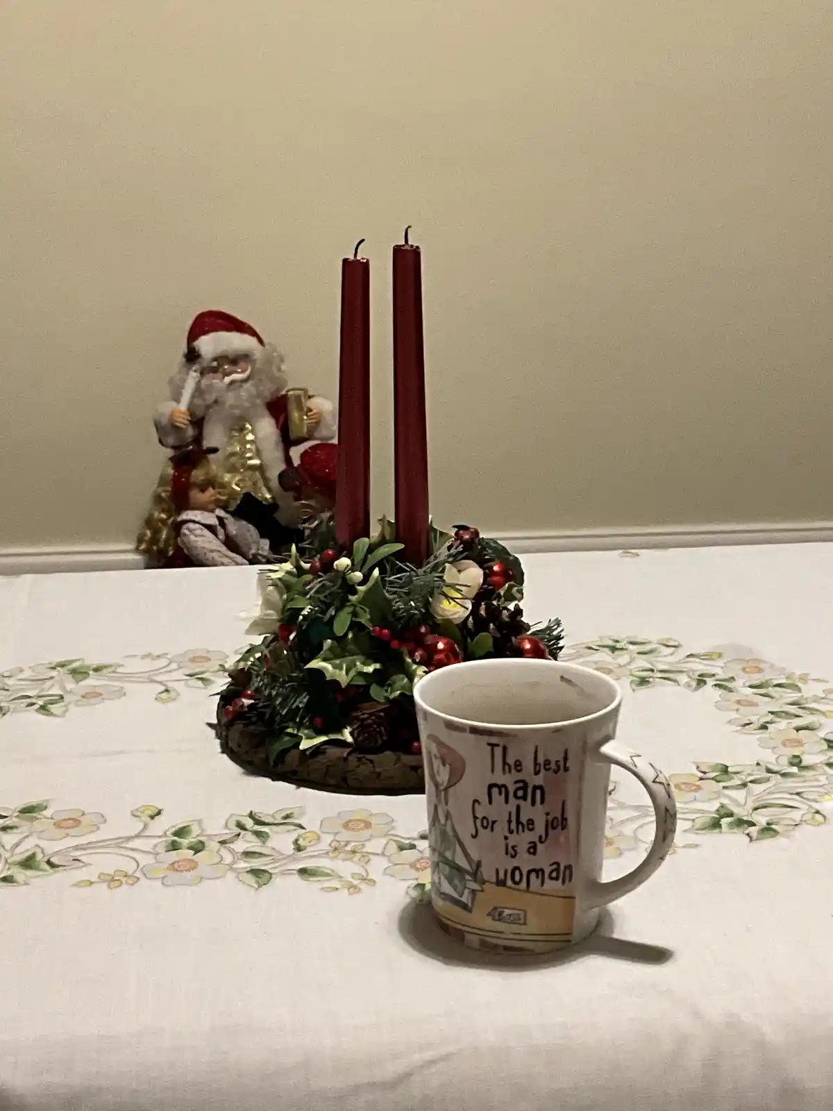 A white ceramic mug with the text 'The best man for the job is a woman' sits on a floral tablecloth, next to a Christmas…