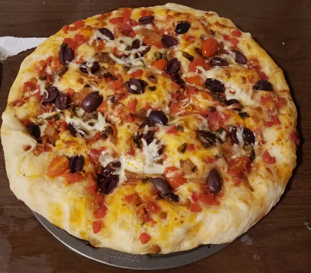 A homemade pizza with melted cheese, black olives, cherry tomatoes, and other toppings sits on a metal baking sheet on a…