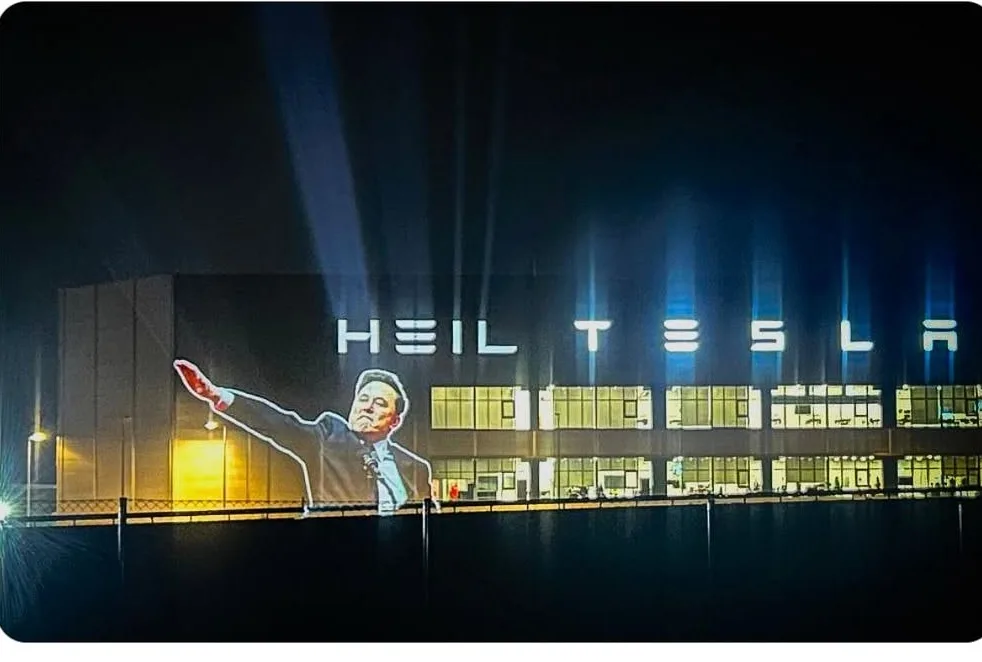 A nighttime photo of a large building with illuminated signage reading 'HEIL TESLA'. Projected onto the side of the building…