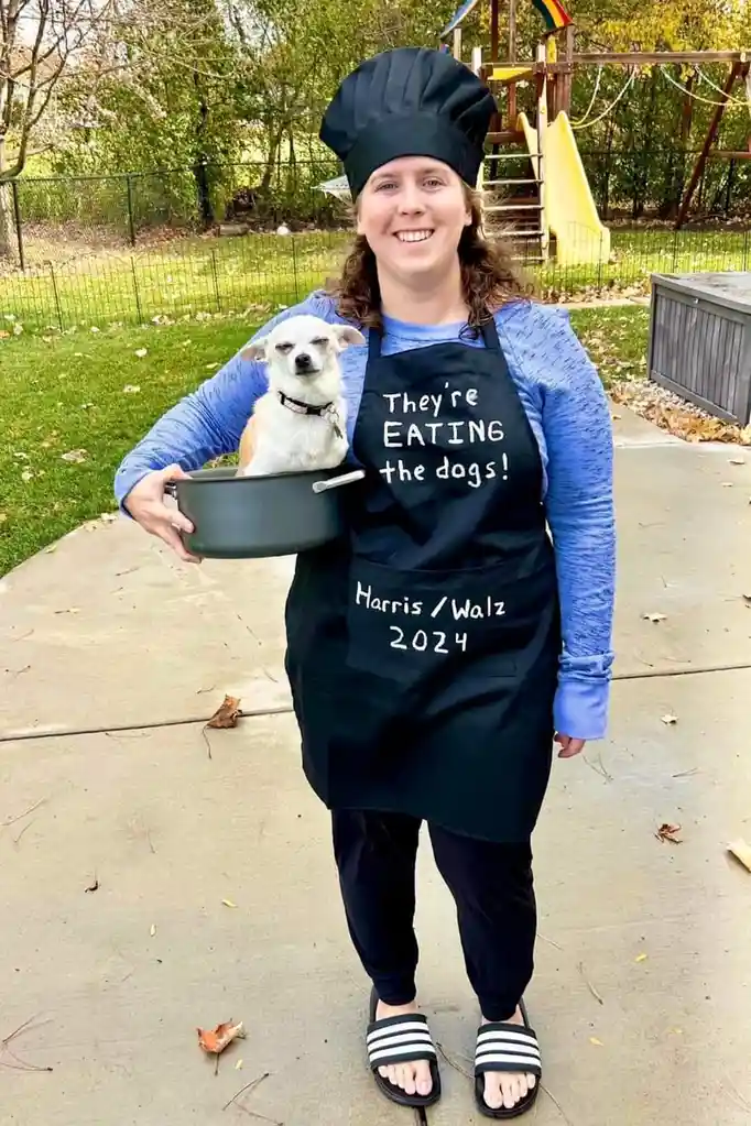 A person wearing a black chef's hat and apron stands outdoors holding a pot with a small dog inside. The apron has white…