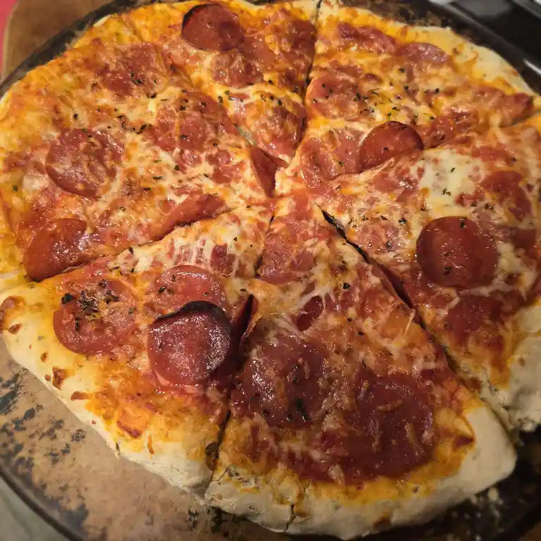 A close-up photo of a pepperoni pizza on a dark baking tray, cut into slices with melted cheese and herbs. One slice in the…