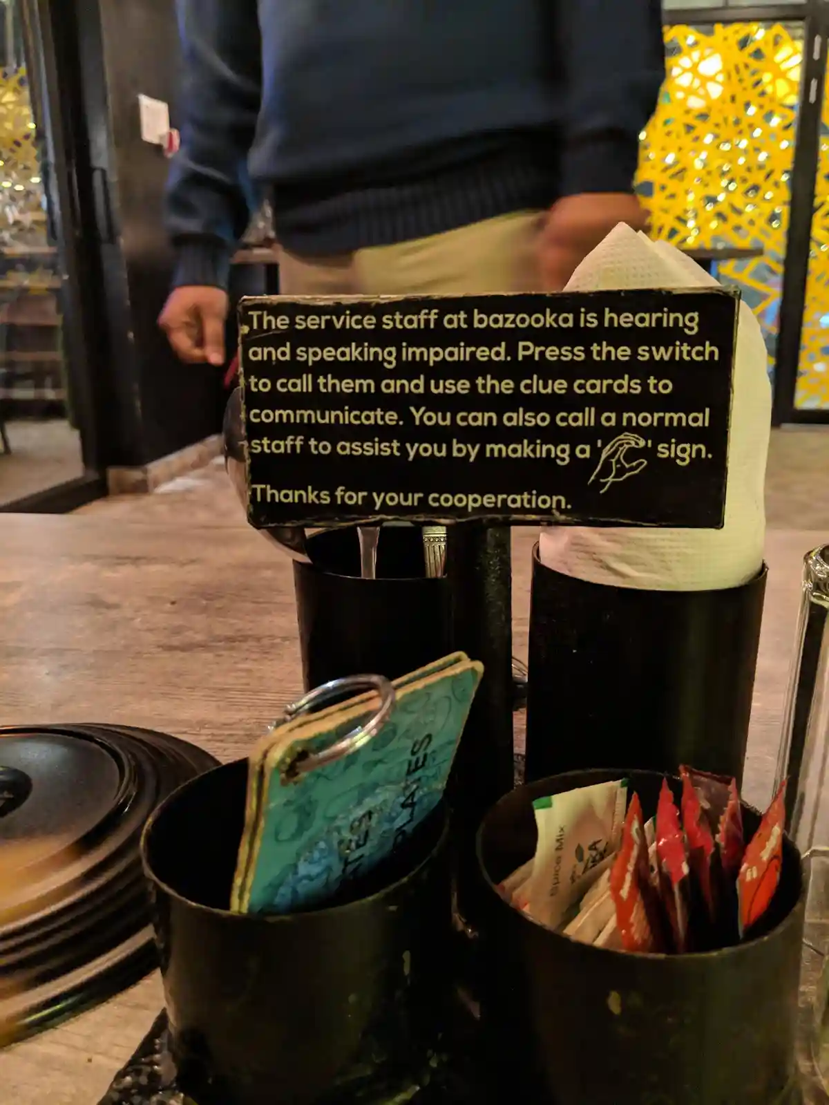 A close-up photo of a restaurant table with condiment holders, a napkin dispenser, and a sign that humorously states the…