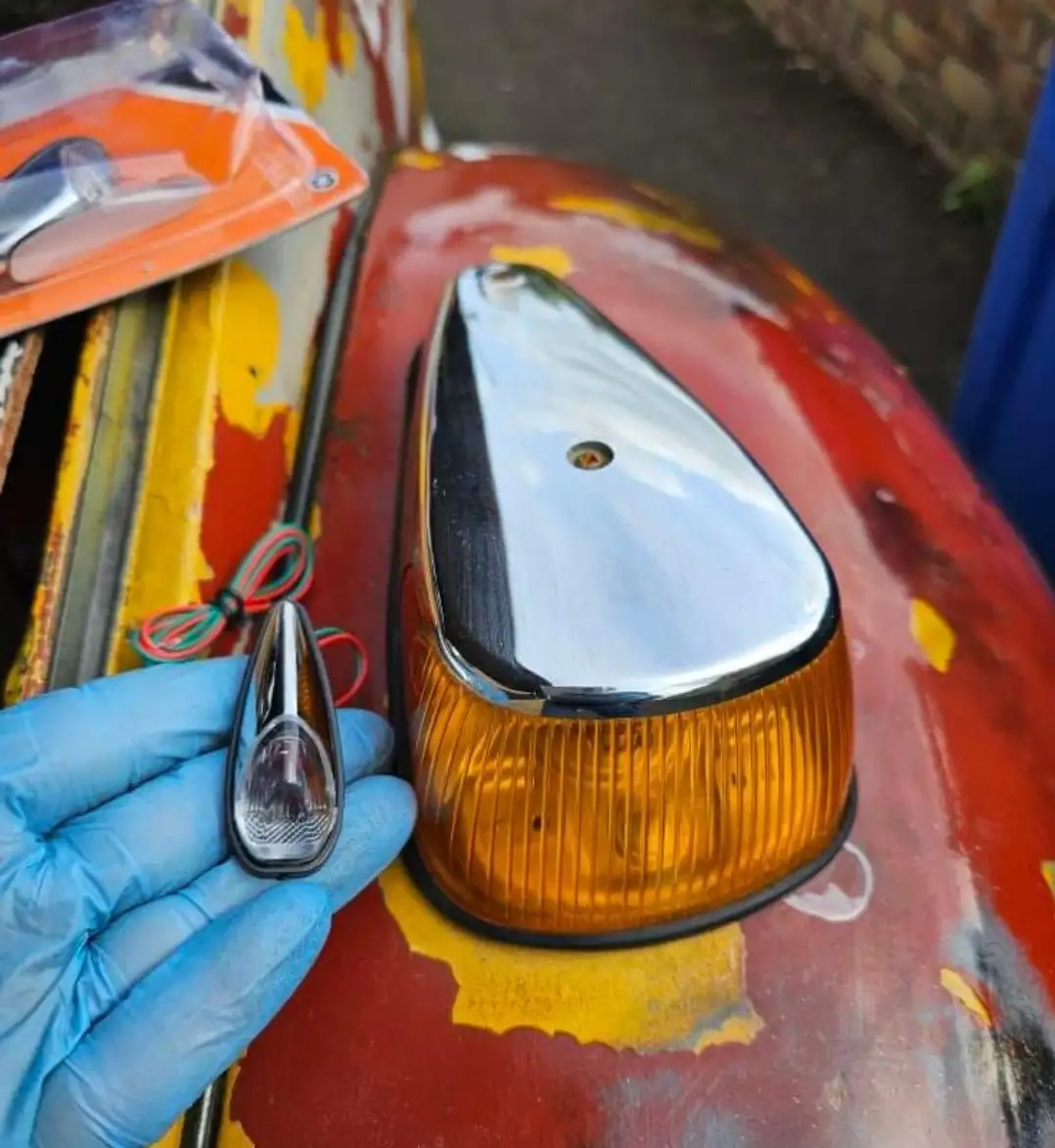 A hand in a blue nitrile glove holds a small, modern, clear LED indicator light next to a large, chrome-topped…