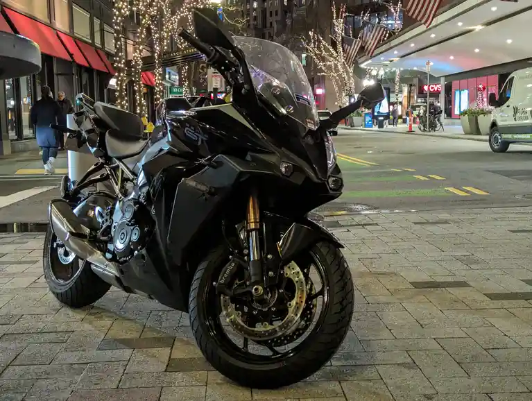 A black sport motorcycle is parked on a city sidewalk at night, with festive lights on trees and storefronts in the…