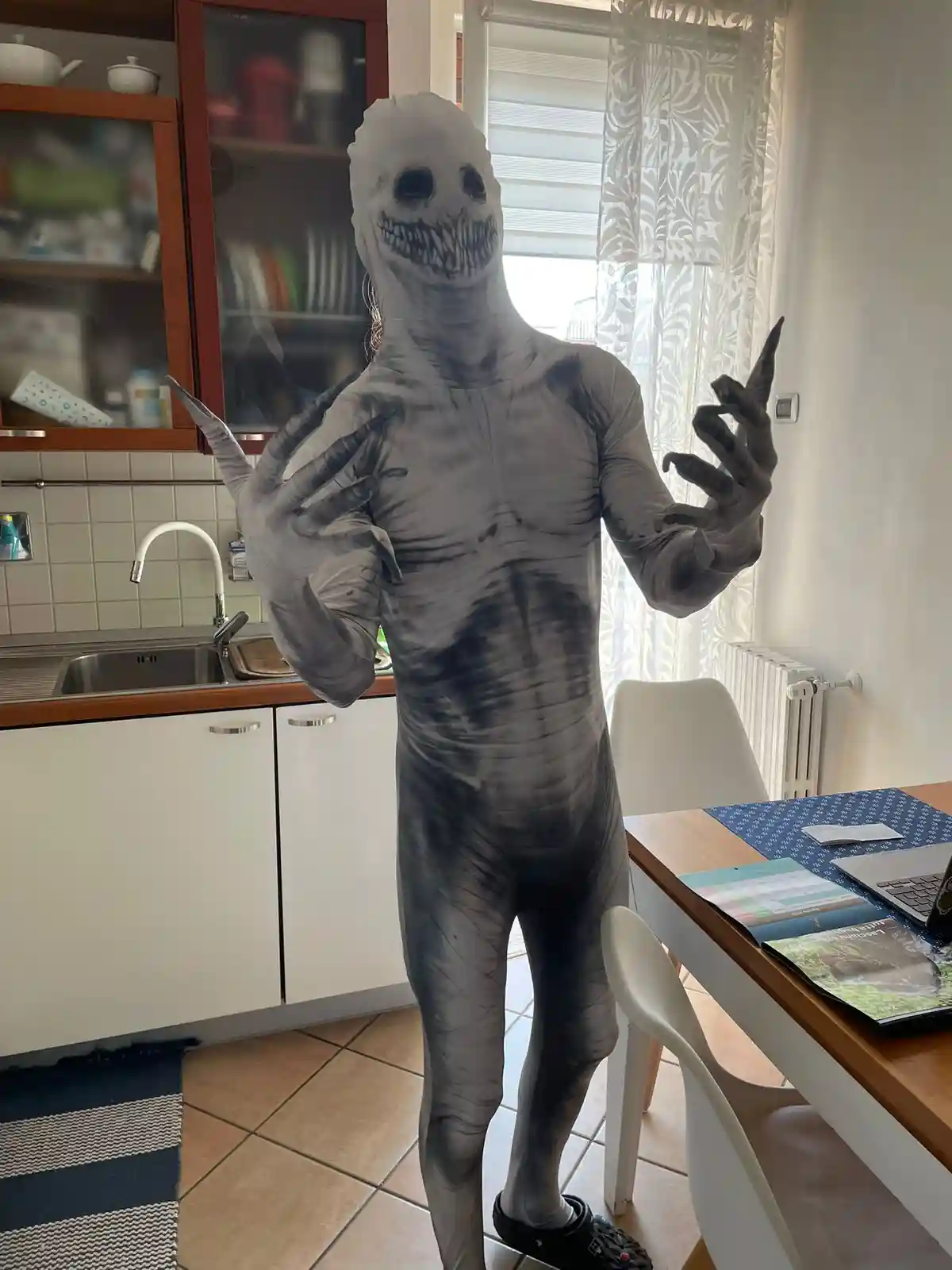 A person in a full-body, grey, textured monster costume with a skull-like face and long, clawed fingers stands in a kitchen…