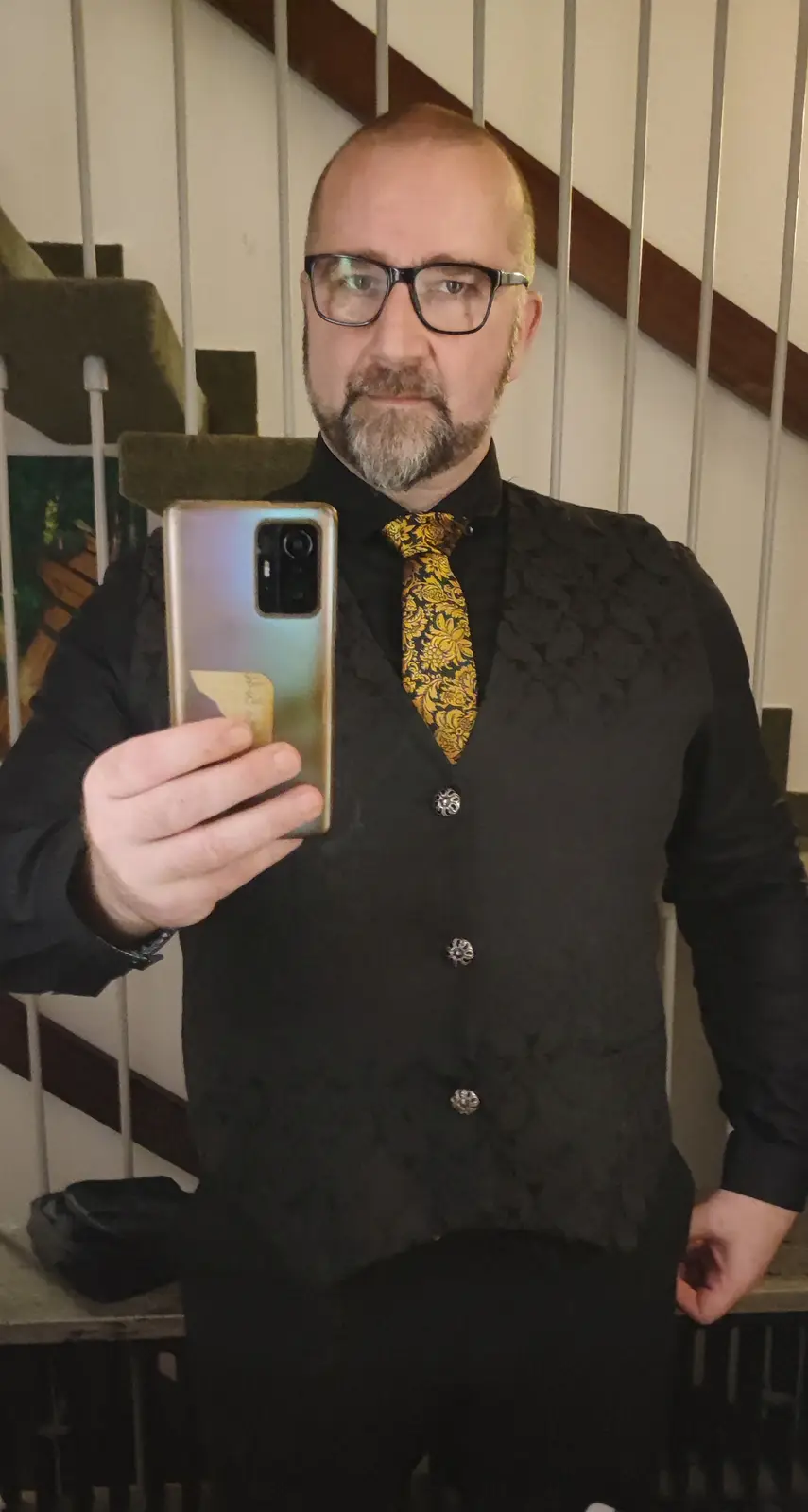 A man with glasses and a beard takes a mirror selfie while wearing a black vest, black shirt, and a bold yellow floral tie…