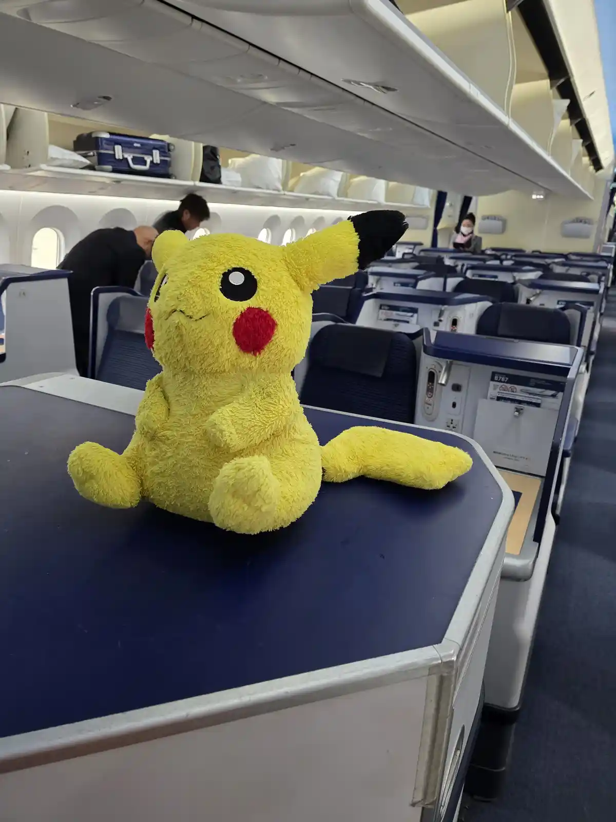 A yellow Pikachu plush toy sits on an airplane tray table with one eye closed and a winking expression, mimicking tiredness…
