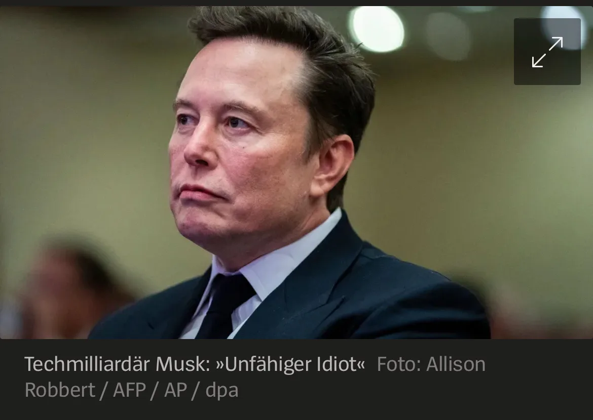 A close-up photo of Elon Musk in a dark suit and tie, looking off to the side with a serious expression. The image has a…