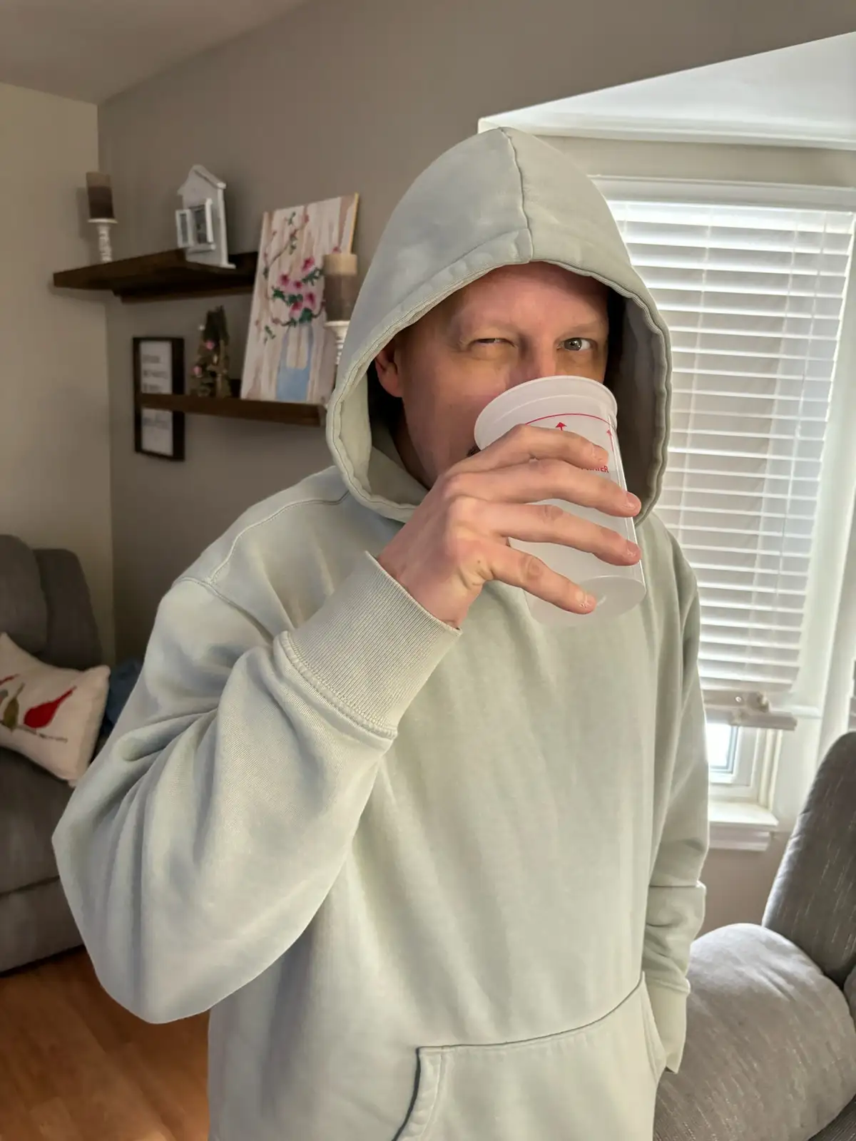 A person in a light-colored hoodie is drinking from a clear plastic measuring cup, looking directly at the camera with a…