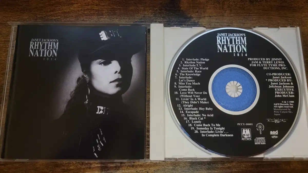 A physical CD case for Janet Jackson's 1989 album 'Rhythm Nation 1814' is open, showing the black-and-white cover art with…