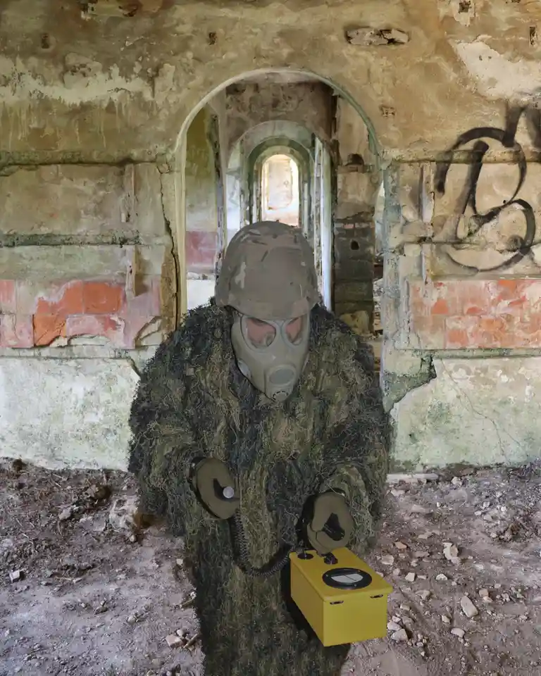 A person in full military gear, including a camouflage helmet and a gas mask, stands inside a dilapidated, crumbling…