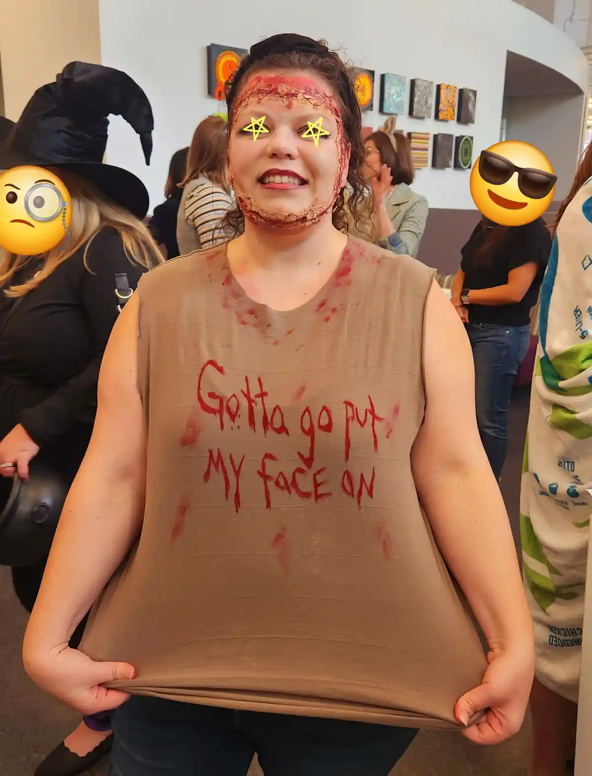 A woman with zombie makeup and a brown tank top that reads 'Gotta go put my face on' in red paint, holding the shirt open to…