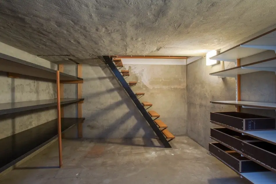 A minimalist, industrial-style basement with exposed concrete walls and ceiling, featuring a modern staircase with wooden…