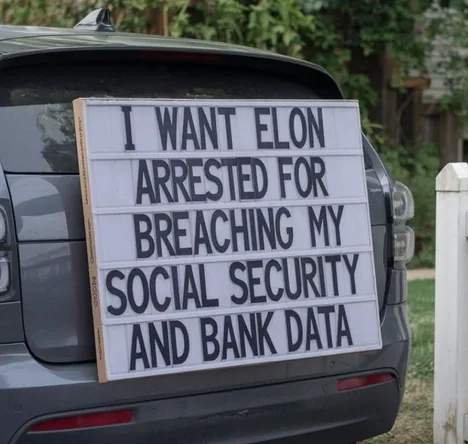 A white lightbox sign with black lettering is mounted on the rear of a dark gray SUV. The sign reads: 'I WANT ELON ARRESTED…
