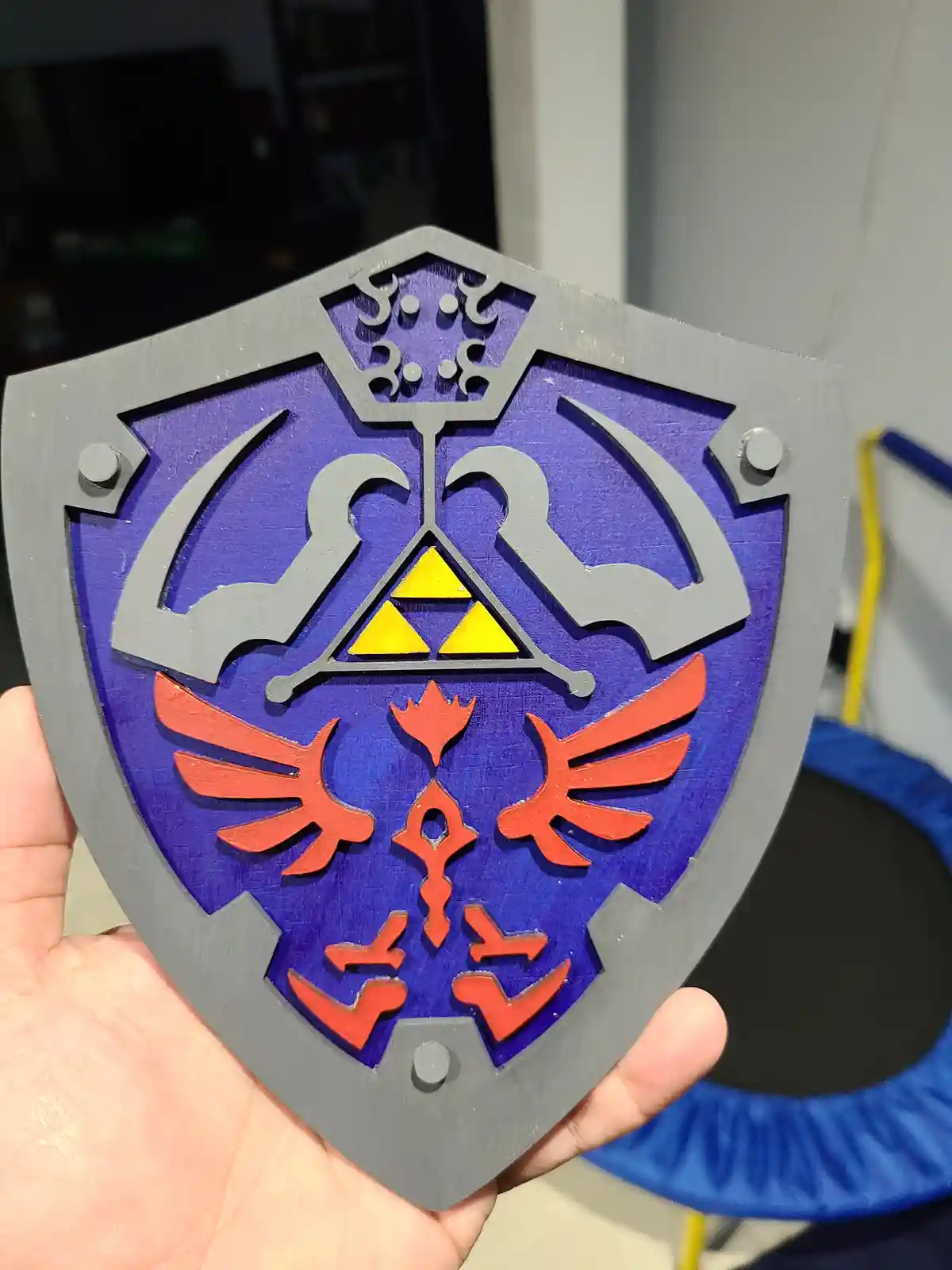A hand holds a 3D-printed replica of the Hylian Shield from The Legend of Zelda, featuring its iconic purple background…