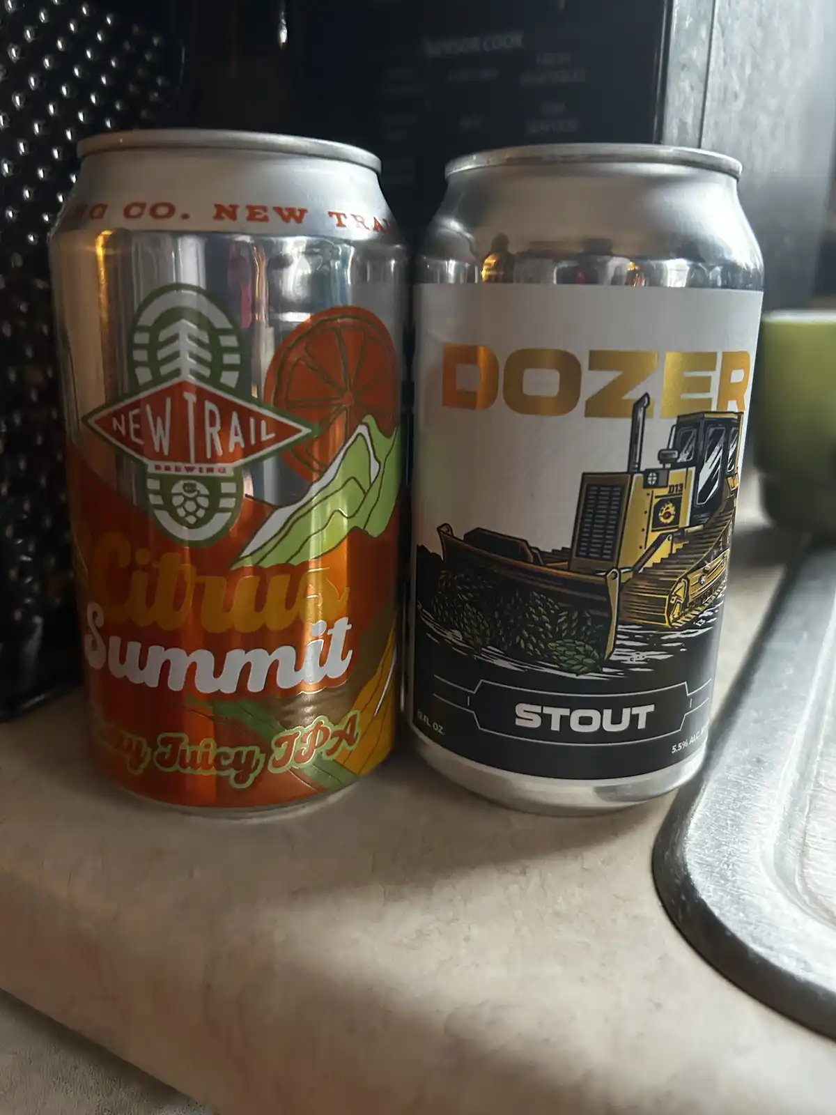 Two beer cans sit side by side on a countertop: a colorful 'Citrus Summit' IPA from New Trail Brewing and a can labeled…