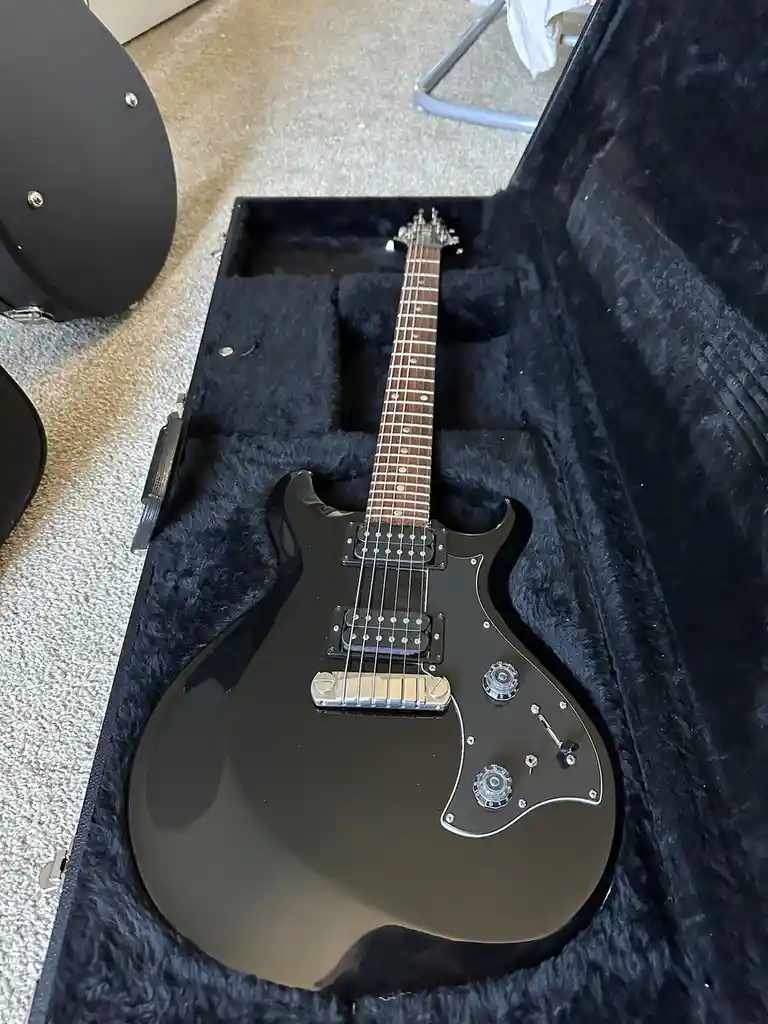 A black electric guitar with a glossy finish rests inside its open, black, plush-lined hard case on a beige carpet. The…