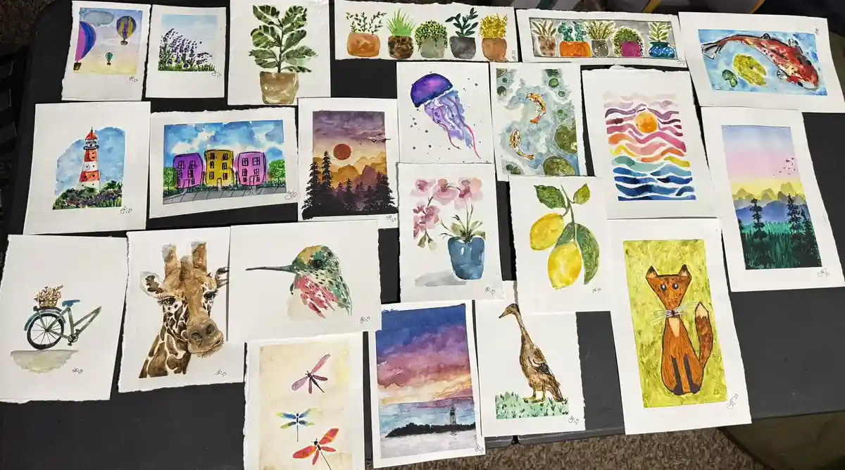 A collection of small watercolor paintings laid out on a dark surface, depicting various subjects including a giraffe, a…