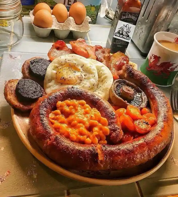 A large plate of a traditional British full English breakfast, featuring fried eggs, bacon, sausages, baked beans, black…