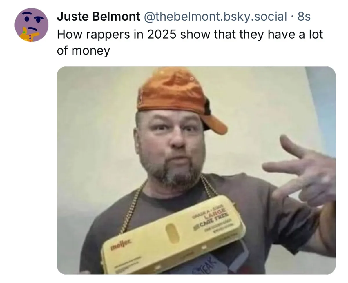 A man wearing an orange cap and a gold chain holds a large yellow egg carton labeled 'moljaz' and 'LARGE CAGE FREE' while…