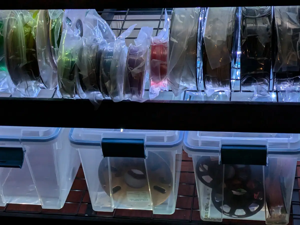 A shelf displays numerous spools of 3D printer filament in clear plastic bags on the top rack, with more filament spools…