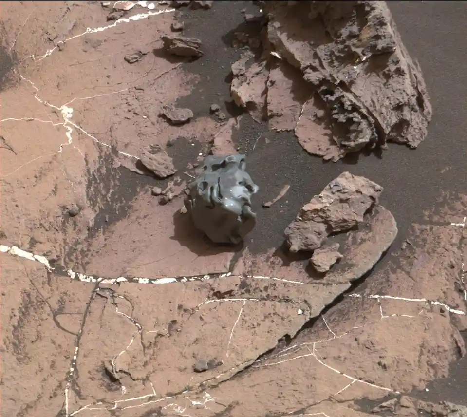 A close-up image of a gray, irregularly shaped rock resting on cracked, reddish-brown Martian soil, with larger rock…