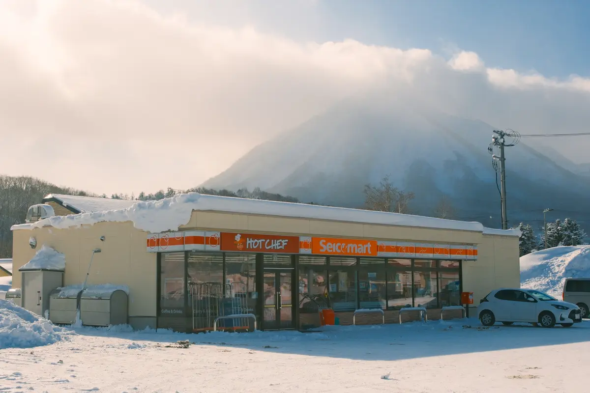 A snowy landscape features a beige convenience store with 'HOT CHEF' and 'Seicomart' signs, set against a backdrop of a…