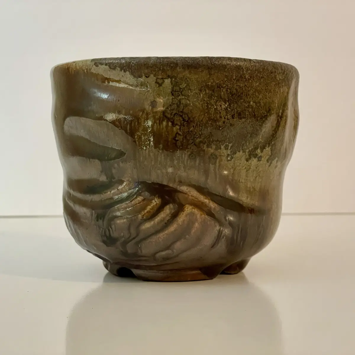 A brown ceramic cup with a glossy glaze sits on a white surface, its body sculpted to resemble a face with a downturned…