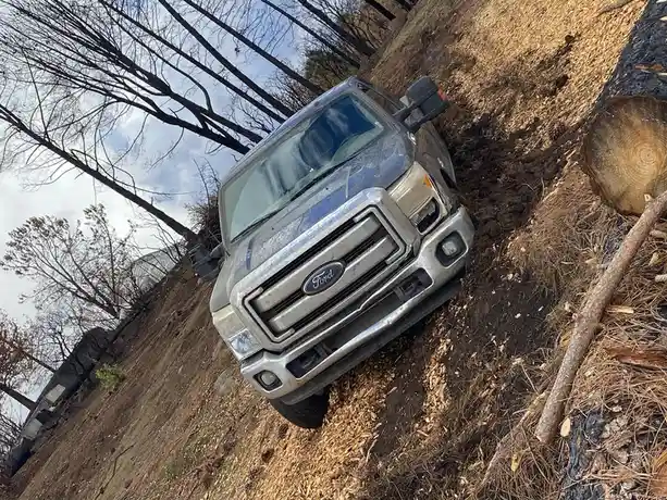 A silver Ford Super Duty pickup truck is angled steeply upward on a dirt and wood-chip slope in a wooded area, with bare…