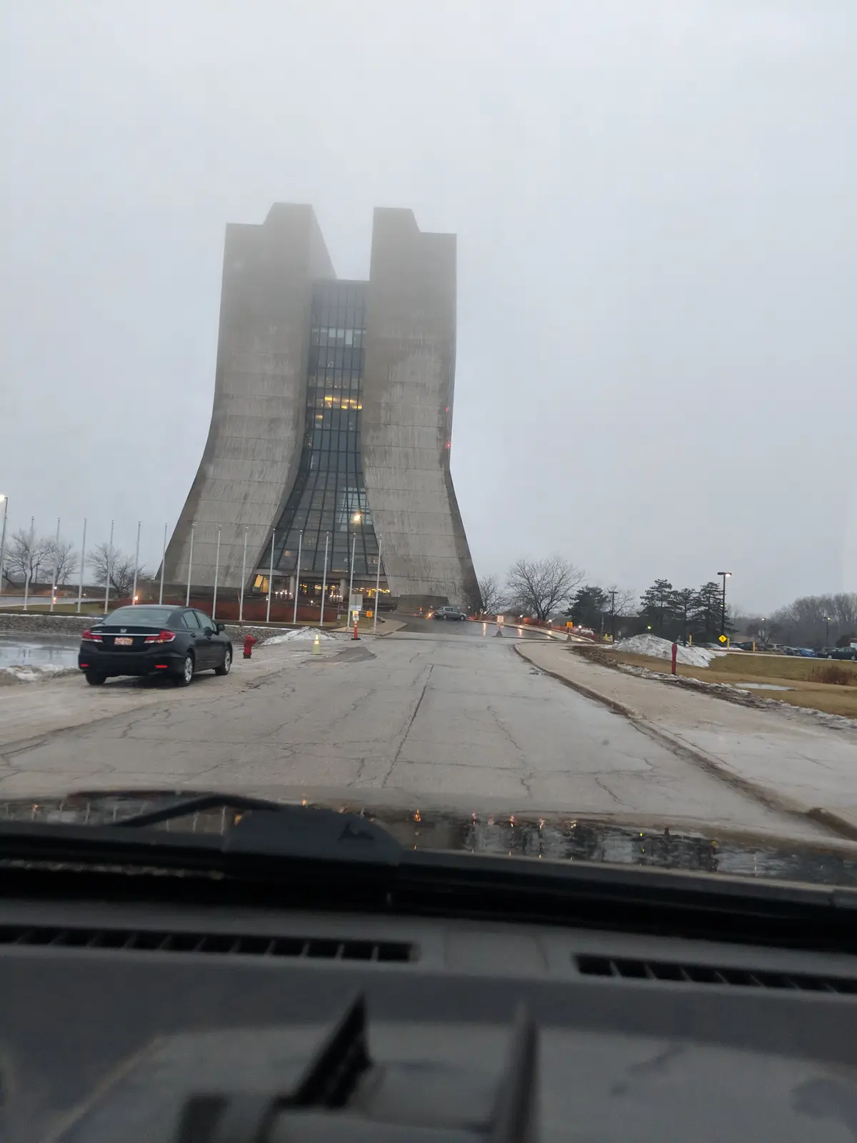 A photo taken from inside a car shows a large, modern concrete building with two tall, curved towers and a glass facade…