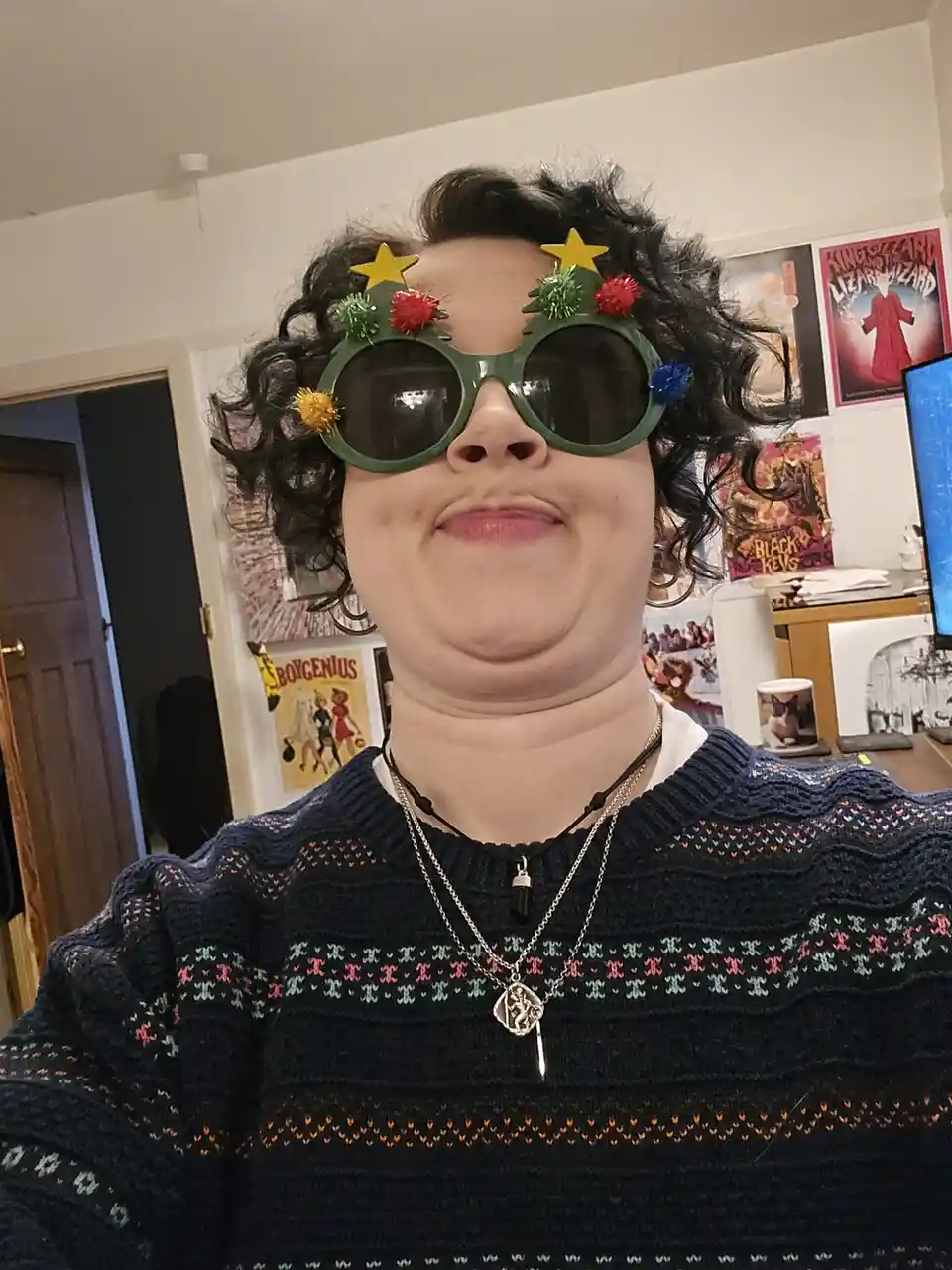 A person with curly dark hair wears oversized green sunglasses decorated with yellow stars, red and green pom-poms, and a…