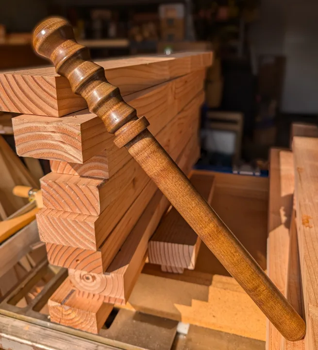 A polished wooden mallet rests diagonally across a stack of freshly cut lumber in a woodworking shop. The background shows…