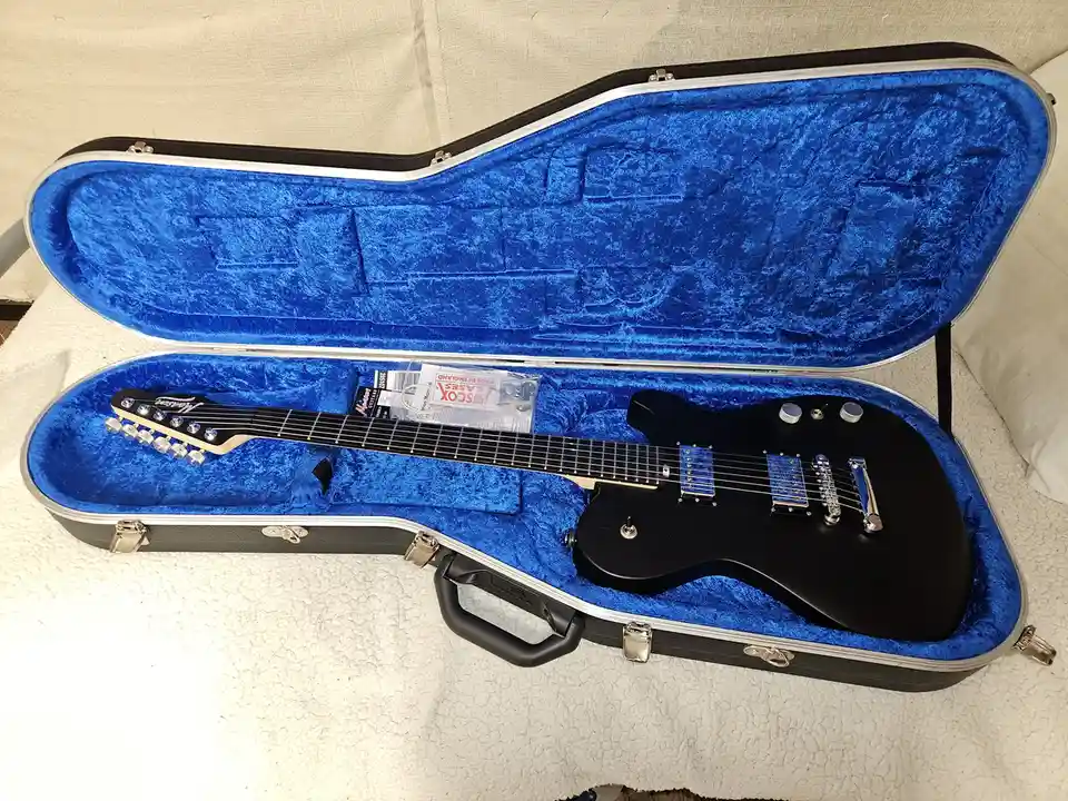 A black electric guitar with a maple neck and blue pickguard rests inside an open, blue velvet-lined hardshell case. The…