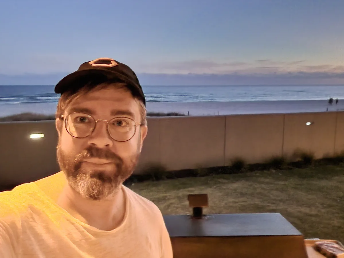 A man with glasses and a beard, wearing a cap and a yellow shirt, takes a selfie at dusk with a beach and ocean in the…