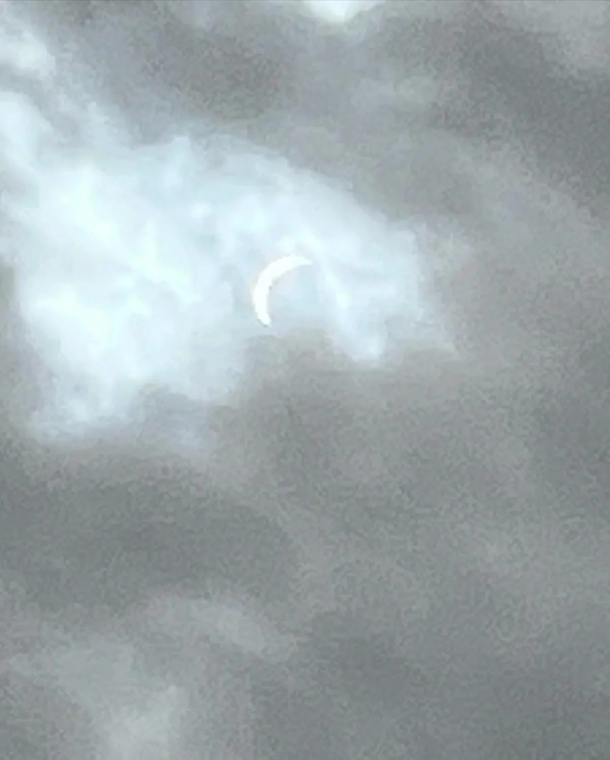 A blurry, low-resolution image shows a bright, crescent-shaped object partially obscured by clouds in a gray sky. The…
