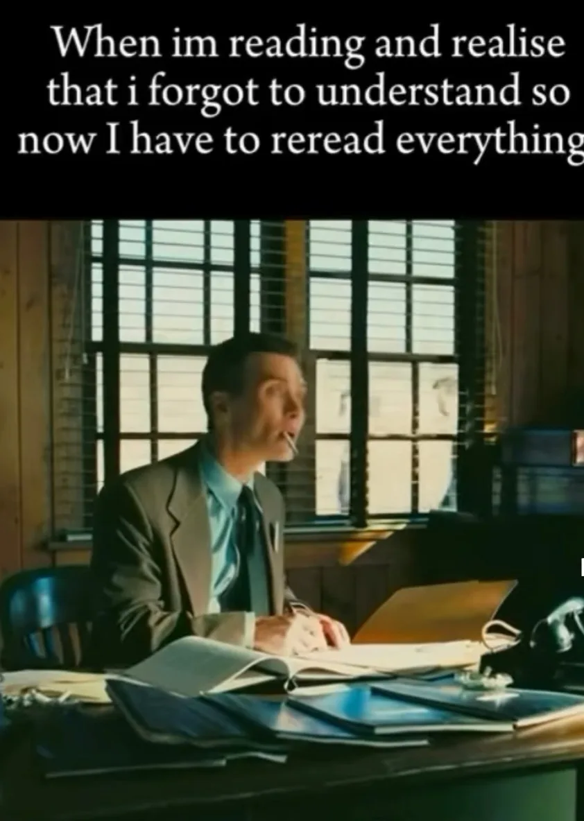 A man in a suit sits at a cluttered desk with open books and papers, looking upward with a shocked expression while holding…