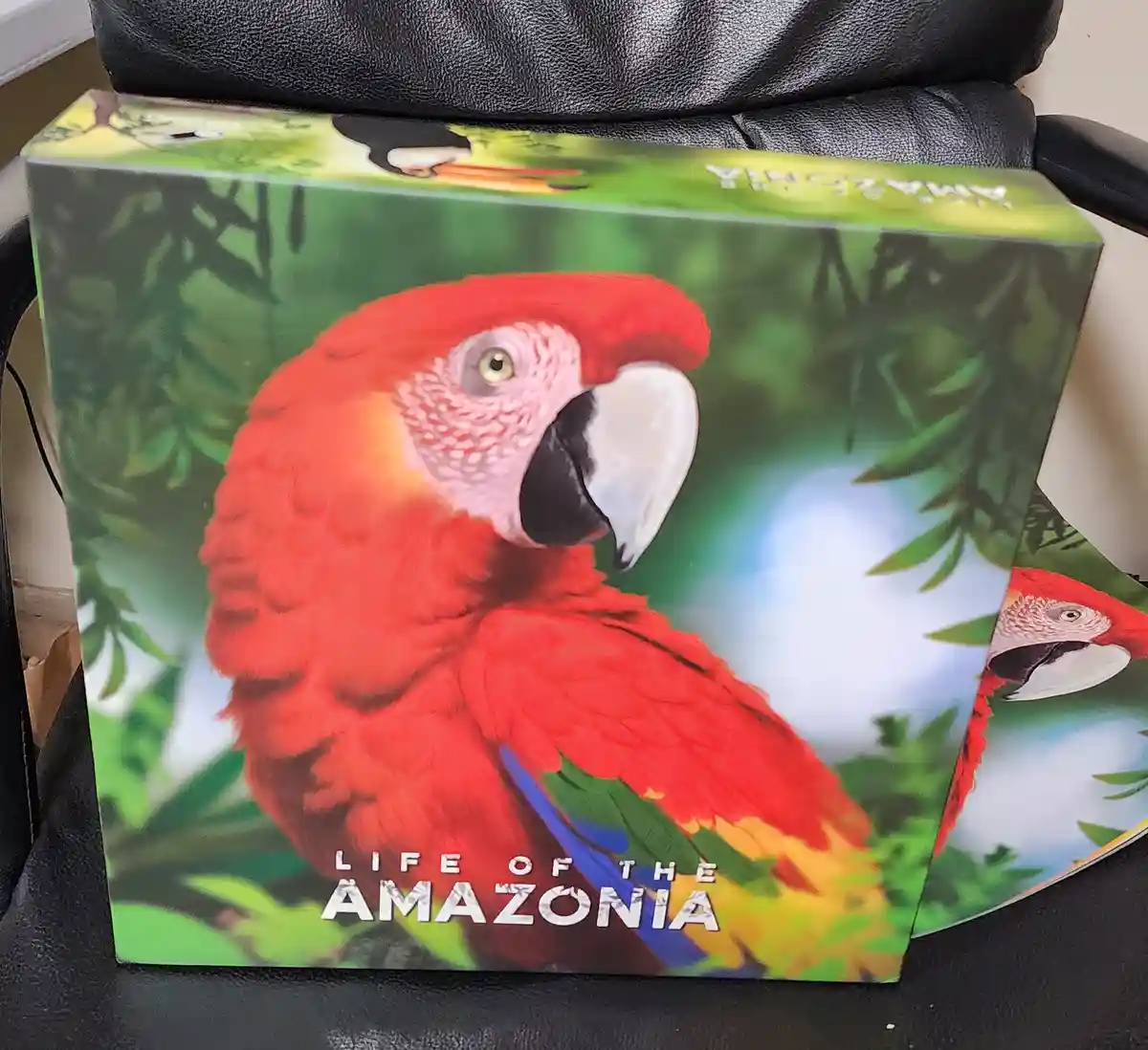 A box for a game or book titled 'Life of the Amazonia' featuring a large, colorful red macaw parrot on its cover, sitting in…