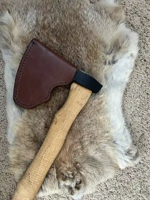 A wooden-handled axe with a brown leather sheath rests diagonally across a large, fluffy animal paw, which appears to be…