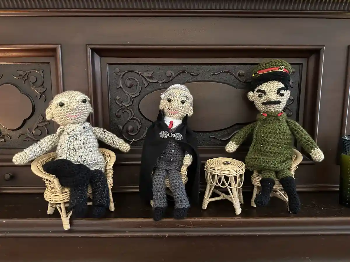 Three crocheted dolls sit on wicker chairs and a stool against a dark wooden panel. The doll on the left is plain, the…
