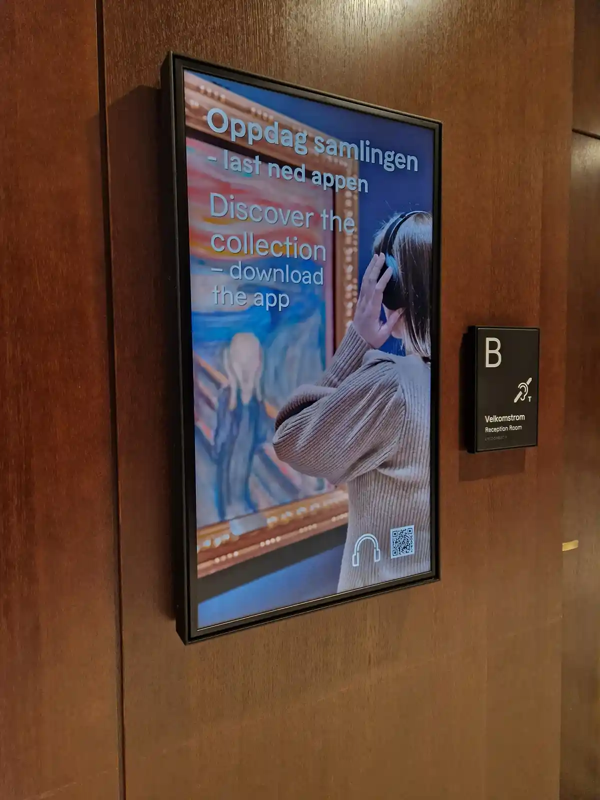 A framed digital poster on a wooden wall advertises an art museum app, showing a person wearing headphones in front of…