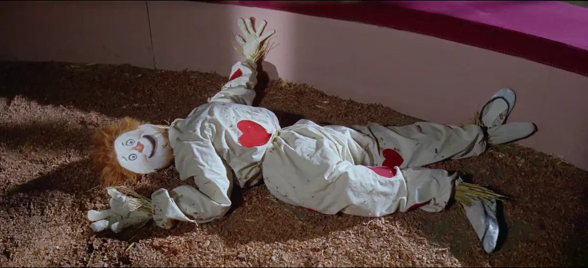 A scarecrow-like figure with a painted face, straw hair, and a white outfit with red heart patches lies sprawled on a bed of…