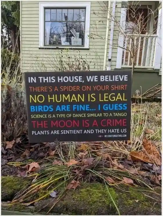 A black yard sign in front of a house lists absurd, satirical beliefs in colorful text, including 'there's a spider on your…