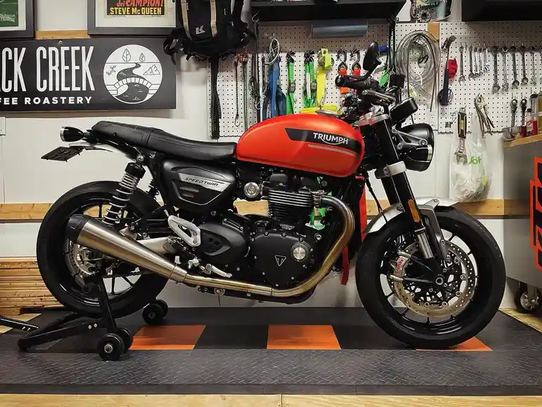 A red and black Triumph Speed Twin motorcycle is parked on a stand in a well-organized garage with tools and a 'Black Creek…