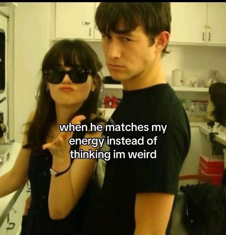A woman wearing sunglasses and making a playful hand gesture stands beside a man in a black t-shirt who looks serious and…