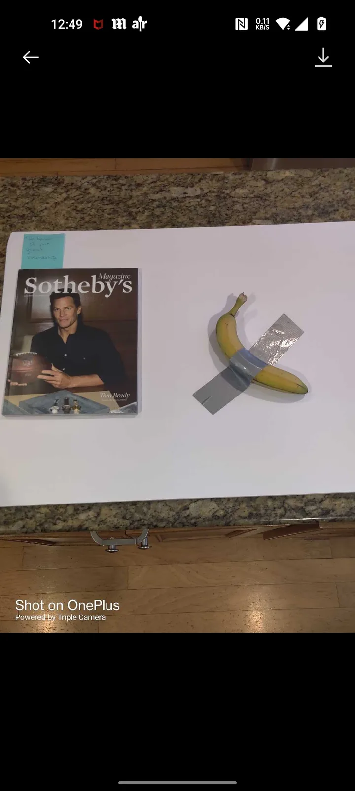 A magazine cover of Sotheby's featuring Tom Brady sits next to a banana taped with duct tape in an X shape on a white…