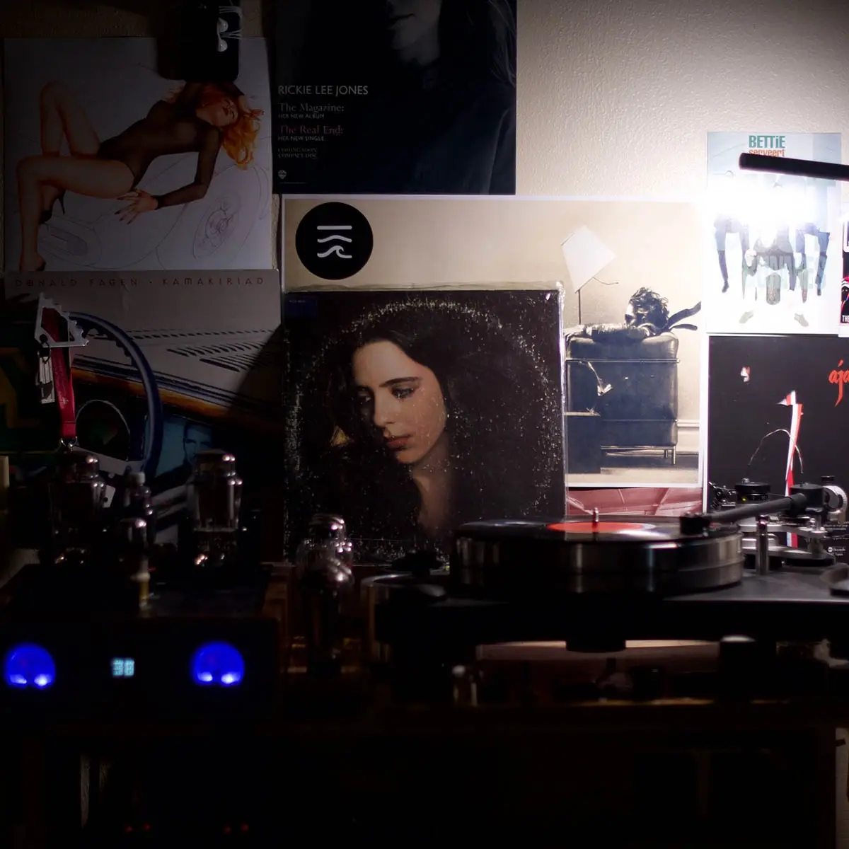 A dimly lit record collection features a vinyl player with a record on the turntable, surrounded by album covers including…