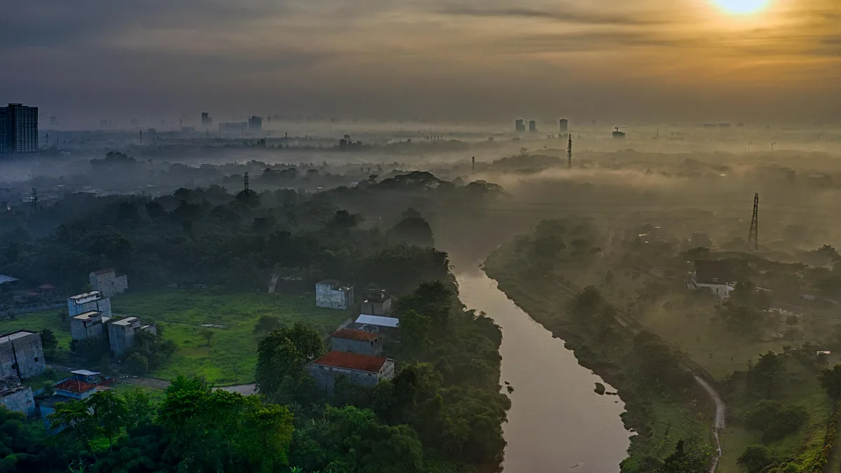 A misty veil shrouds the morning landscape as a sinuous river weaves through a tapestry of emerald greenery, its gentle flow…