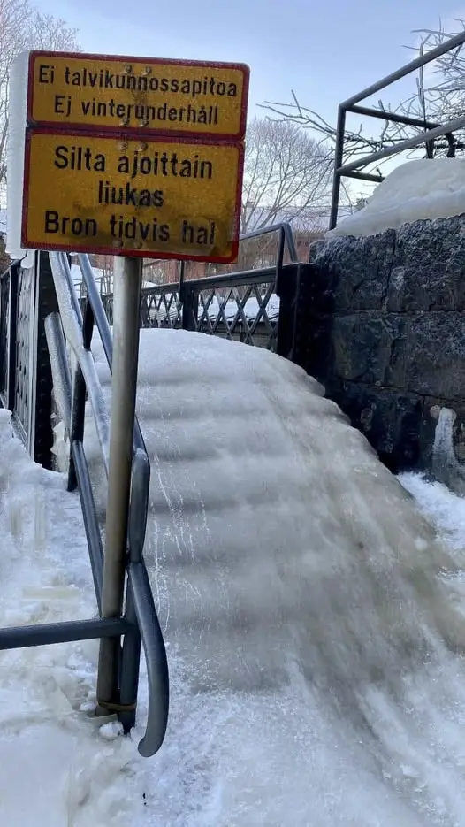 A yellow warning sign in Finnish and Swedish stands beside a snow-covered, icy bridge ramp. The sign warns that the bridge…